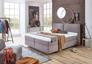 Graues Boxspringbett mit gepolstertem Kopfteil, weißer Matratze und Kissen in Grau und Rostorange in einem hellen Schlafzimmer mit Holzlamellenwand und großen Fenstern